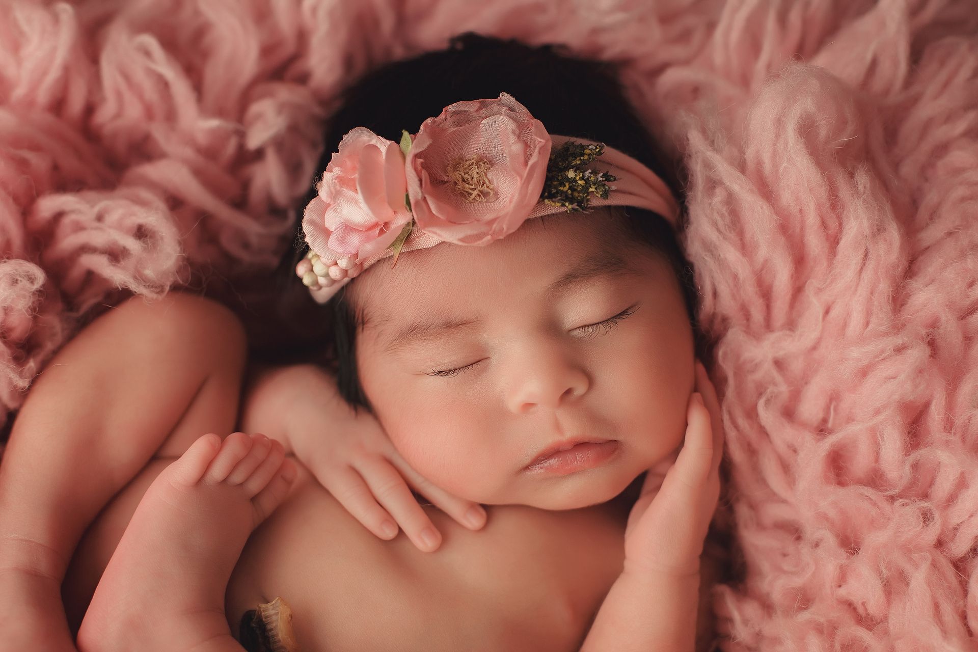 A newborn baby girl wearing a pink flower headband is sleeping on a pink blanket.