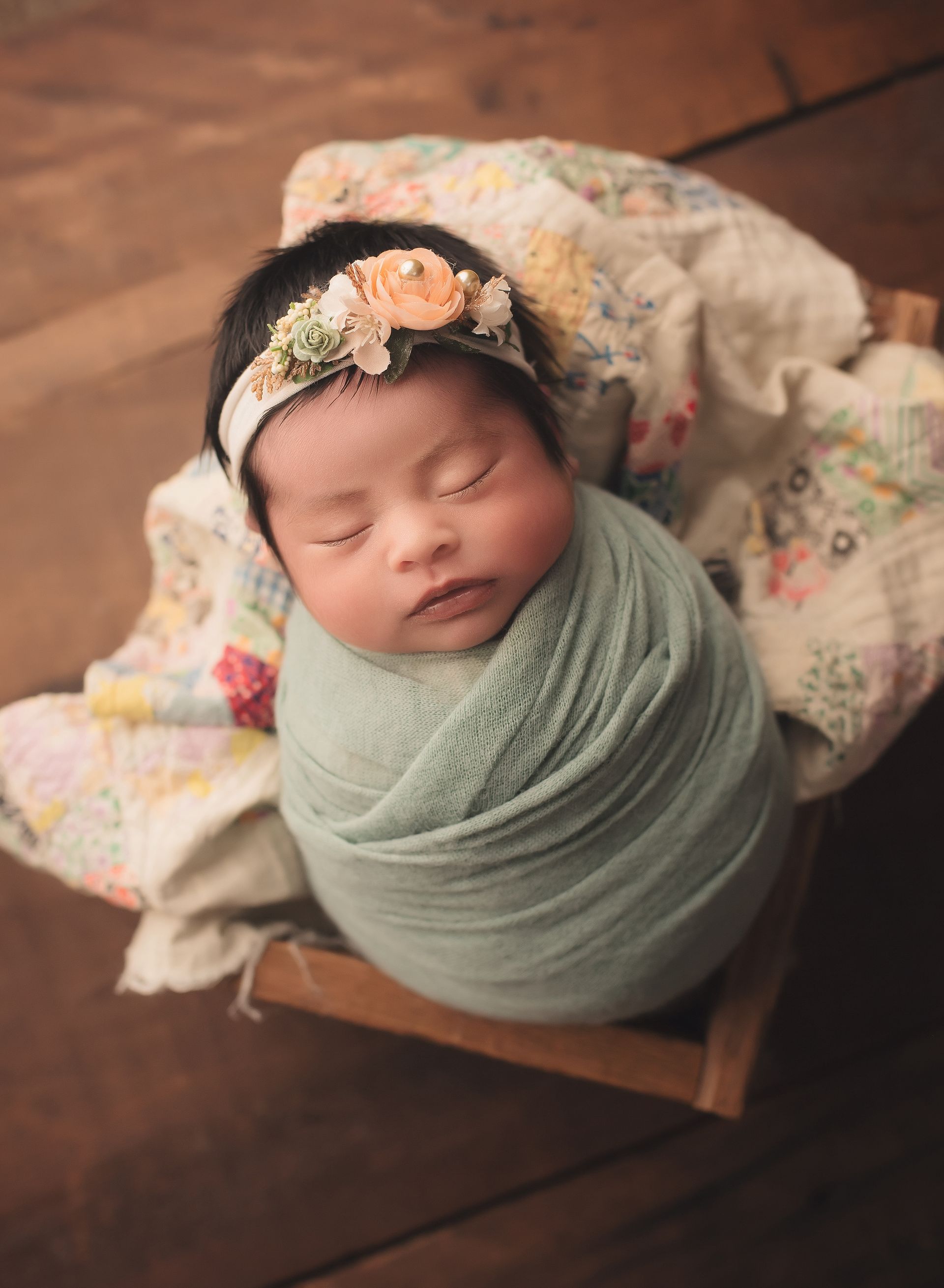 A newborn baby is wrapped in a green blanket and wearing a flower headband.