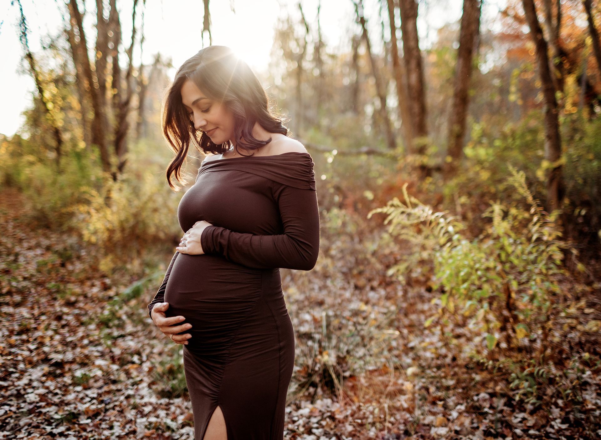 Pregnant woman in brown dress, holding her belly, smiles in a sunlit forest.