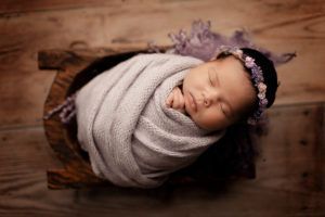 A newborn baby wrapped in a blanket is sleeping in a wooden bucket.