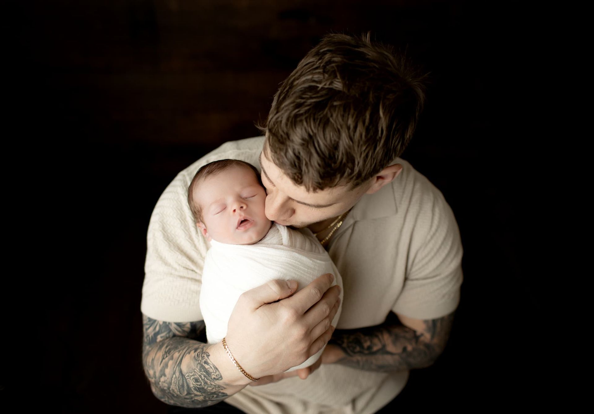 Man holding and kissing a swaddled baby. Dark background. The man has tattoos at a photo session near Bergen County, NJ.