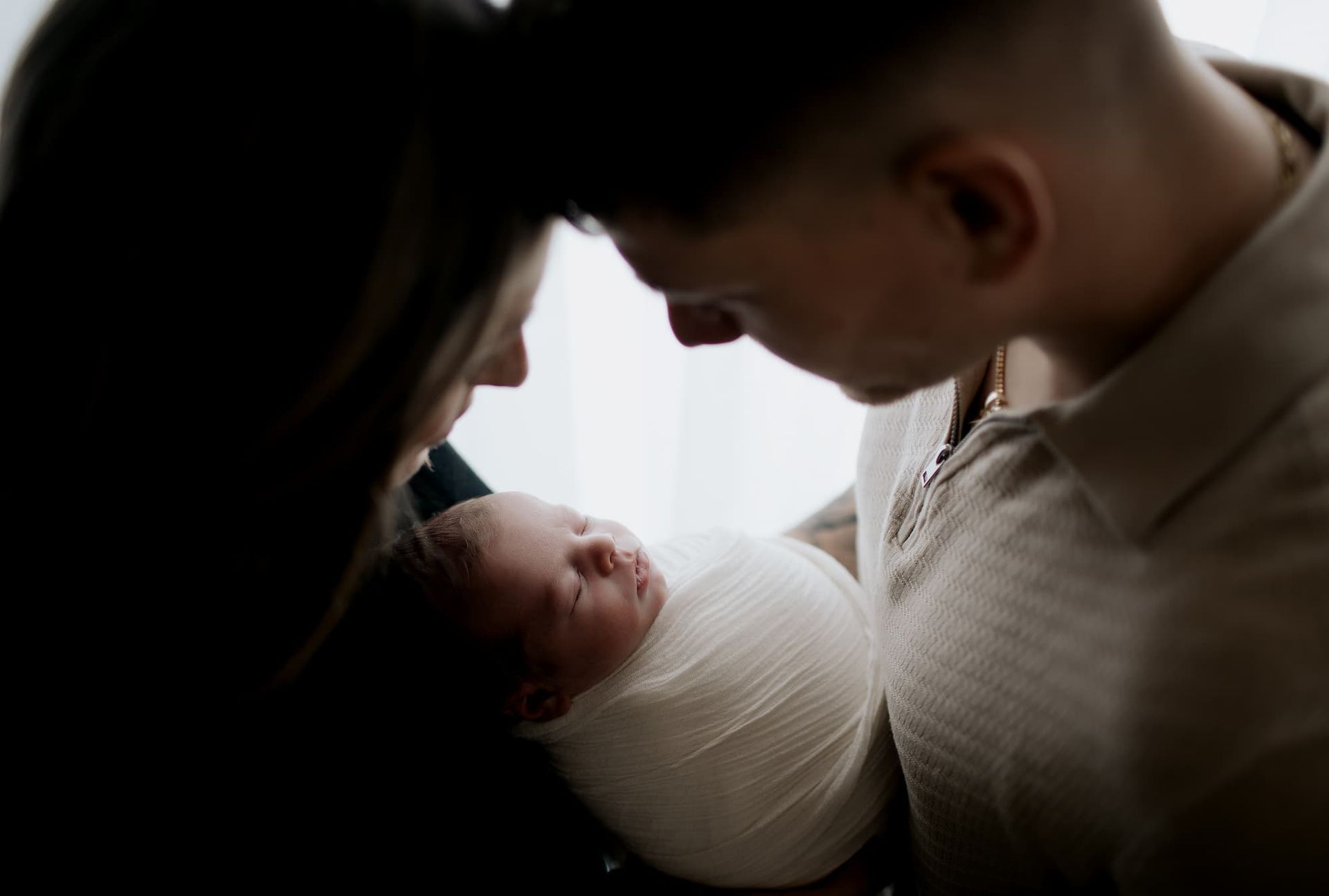 Parents gaze adoringly at newborn baby wrapped in white swaddle at a photoshoot near me.