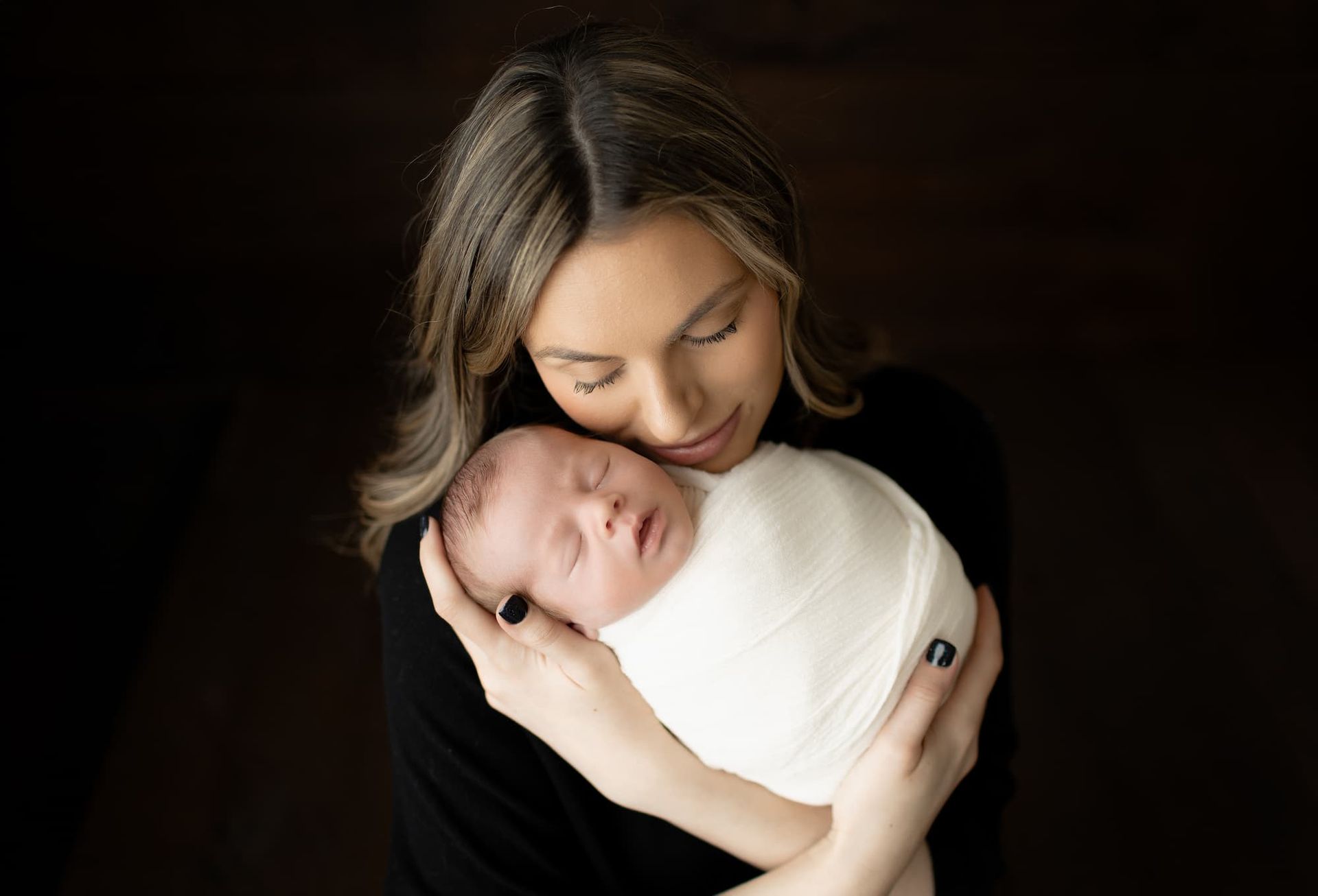 Woman in black holds a swaddled infant, looking down with a gentle expression. Dark background at photoshoot in Bergen County, NJ.