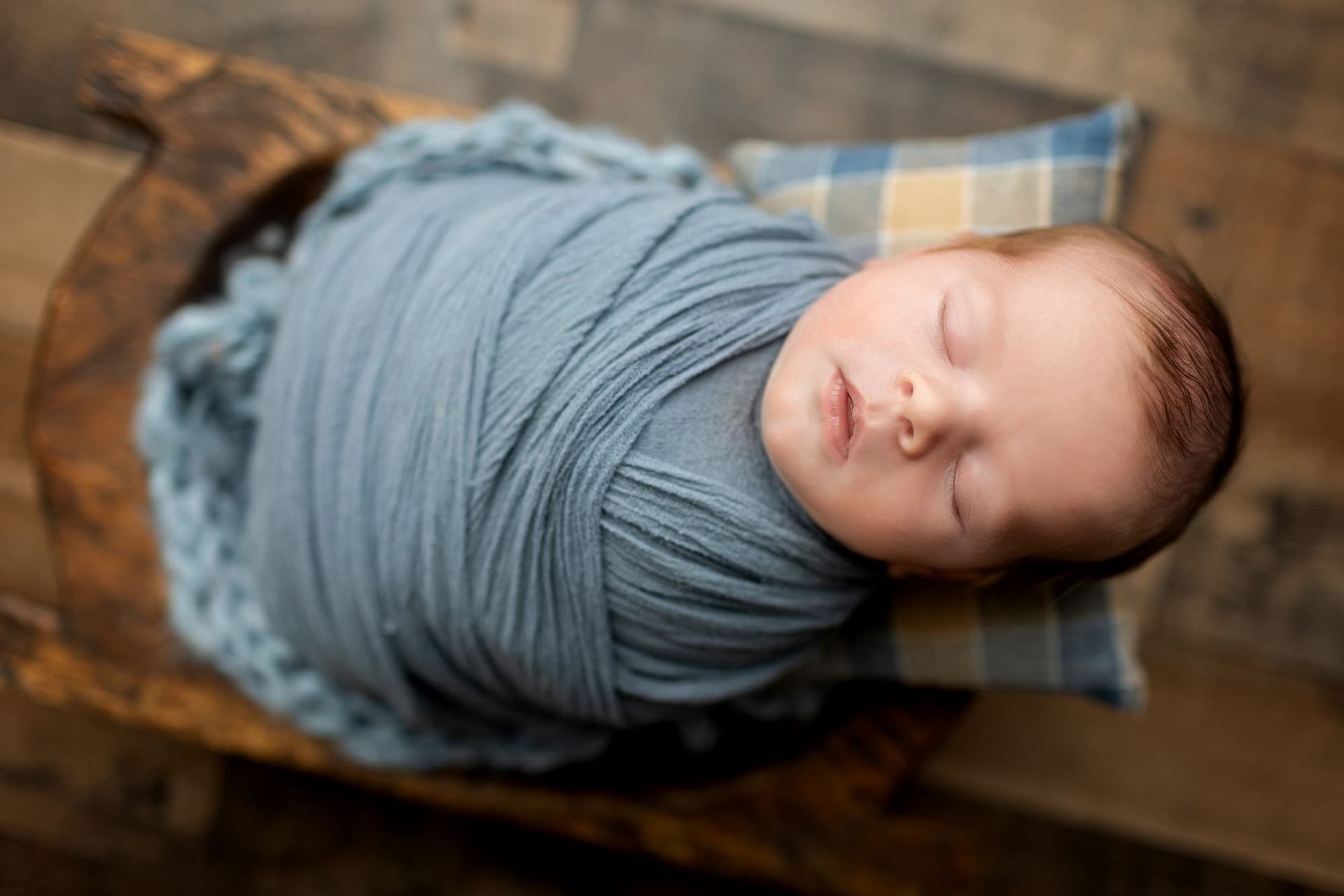 Newborn baby swaddled in blue fabric, sleeping in wooden bowl with small patterned pillow in Bergen County, NJ.