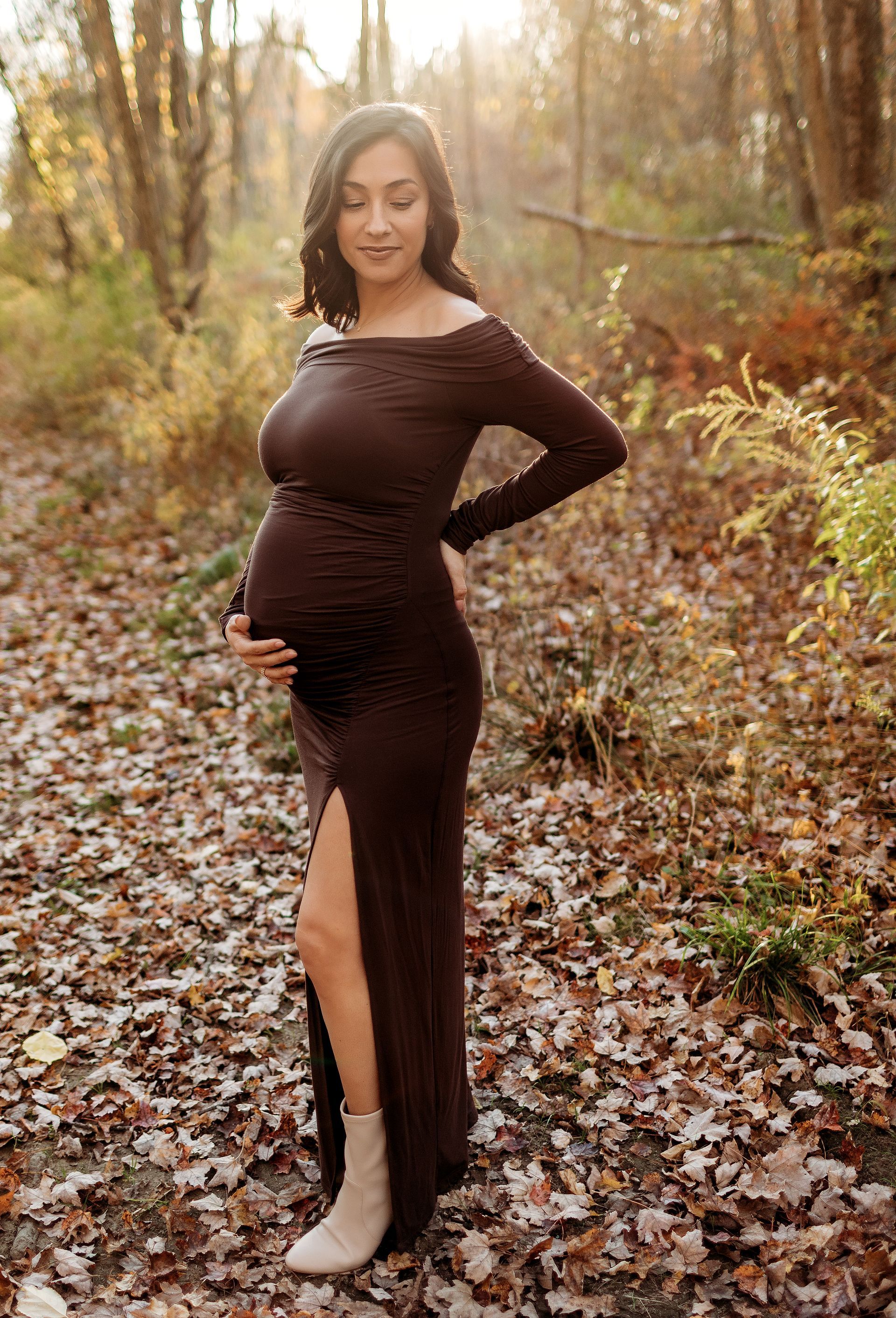 Pregnant woman in brown dress, holding belly, standing in woods with fallen leaves.