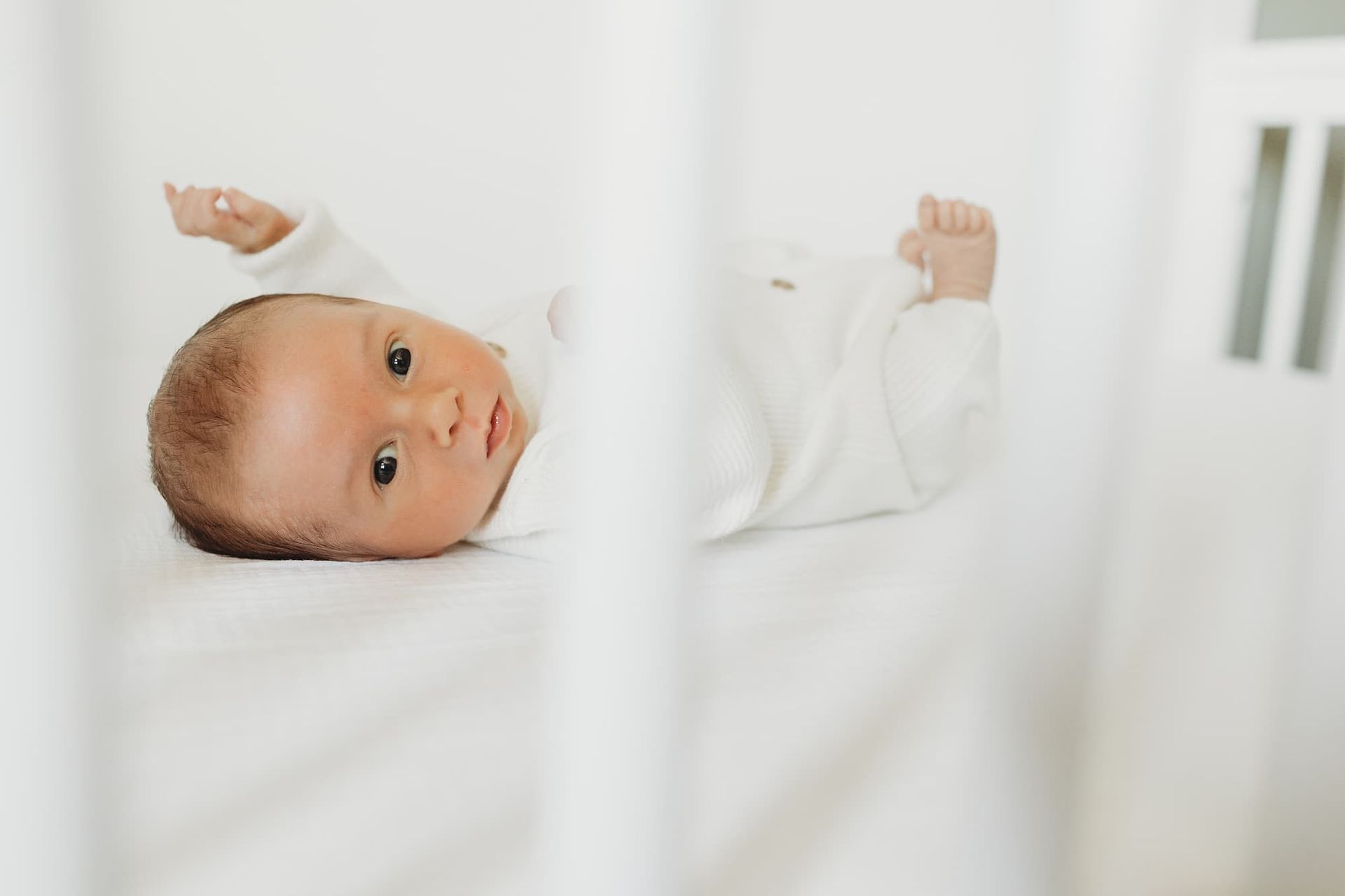 Baby lying in a white crib, looking at the camera with arms raised