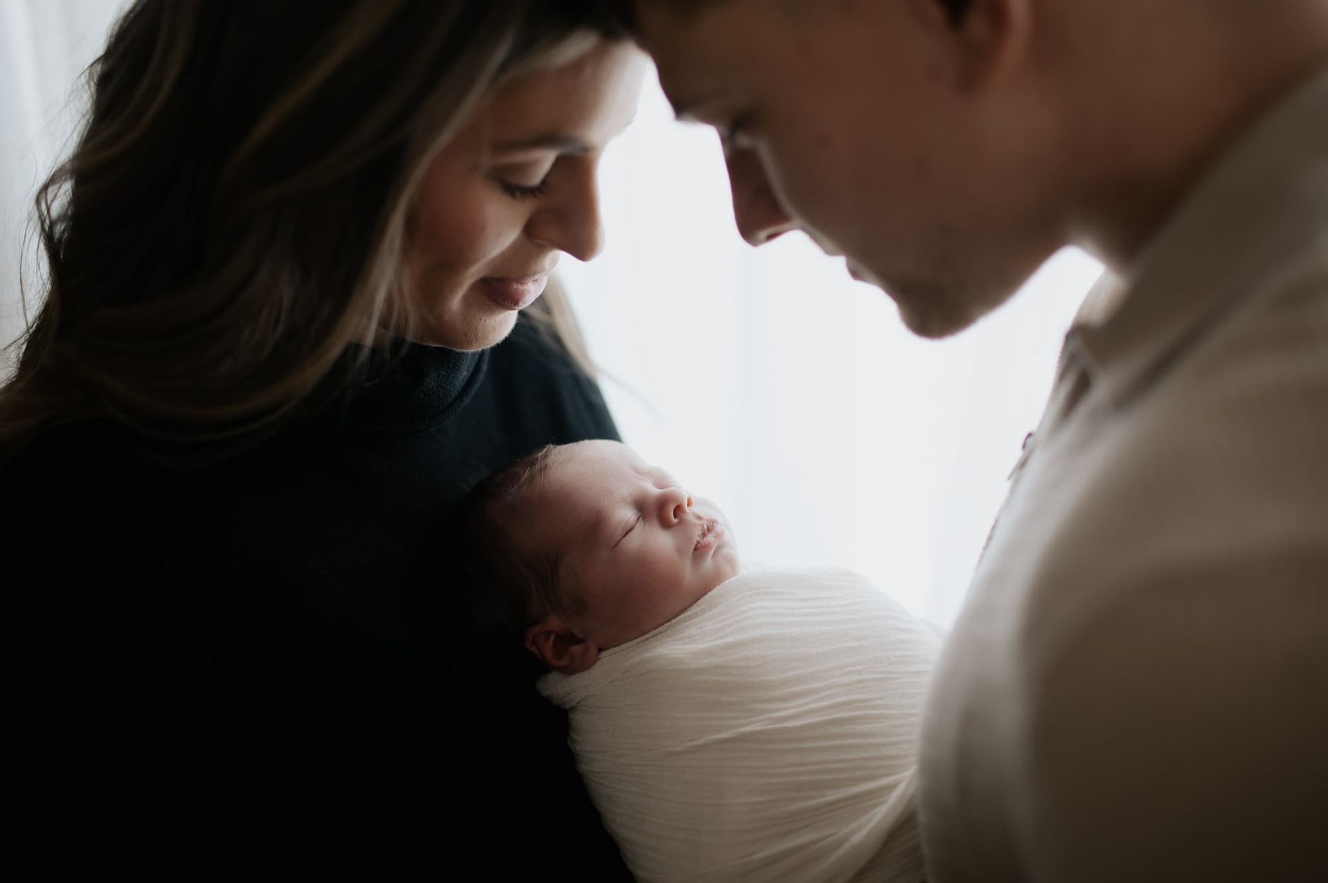 Parents gazing at a swaddled newborn baby, indoors near a window at a photoshoot near Bergen County, NJ.