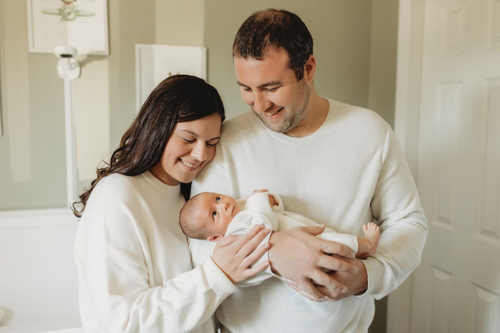 Parents gently hold a newborn in a bright home doorway, smiling down at the baby in Ridgewood, NJ.