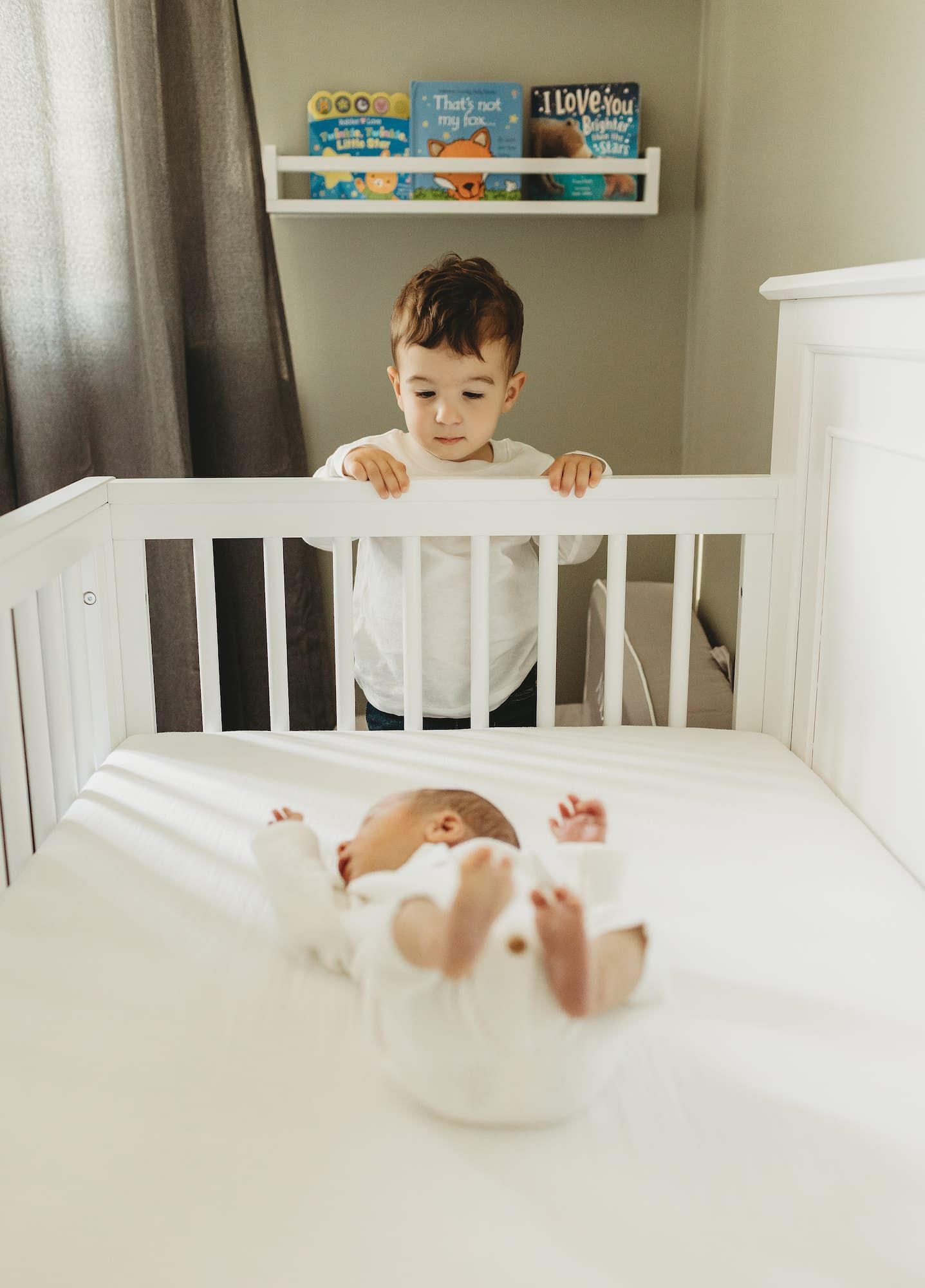 Child peeking over a white crib at a baby lying inside, in a bright nursery with books on the shelf in Ridgewood, NJ.