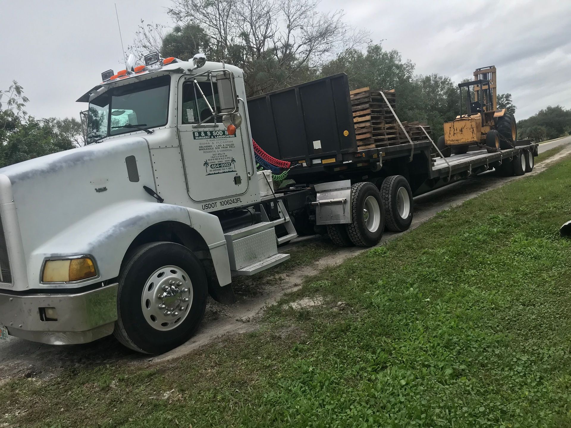 A white semi truck is towing a dumpster and a forklift.