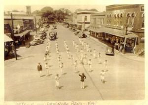 Band marching down a street in Gainesville, Texas, 1941, flanked by parked cars and storefronts.