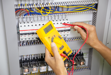 Electrician wearing a yellow hard hat and gloves working on a wall panel near a window.