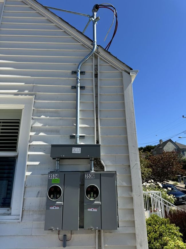 Electrical meter boxes mounted on the side of a white house with conduit leading up to power lines.