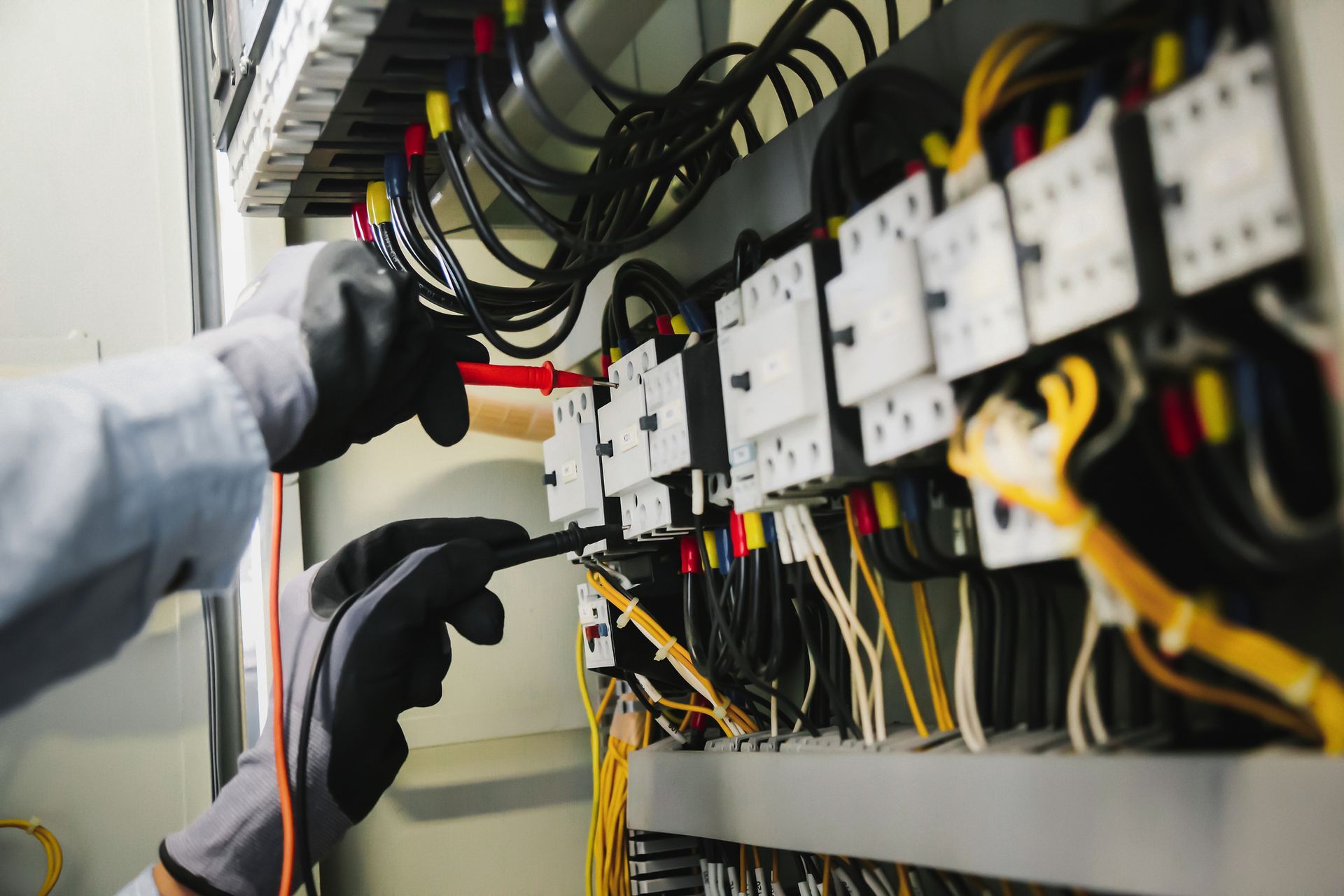 Electrician with gloved hands using a multimeter to test wires inside an electrical panel.