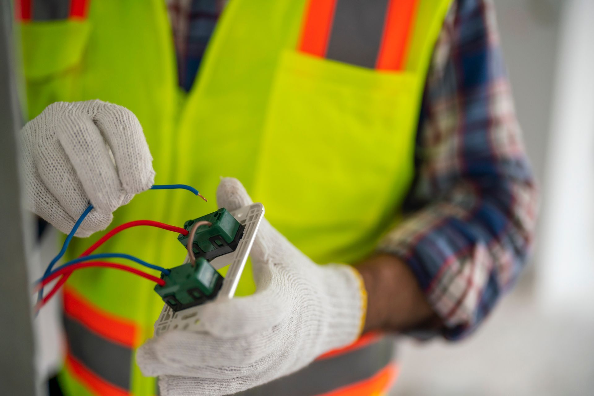 Electrician installing electrical outlet, wearing gloves and safety vest.