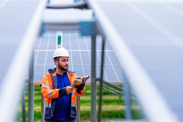 Man in safety vest and hardhat inspecting solar panels, outdoors.