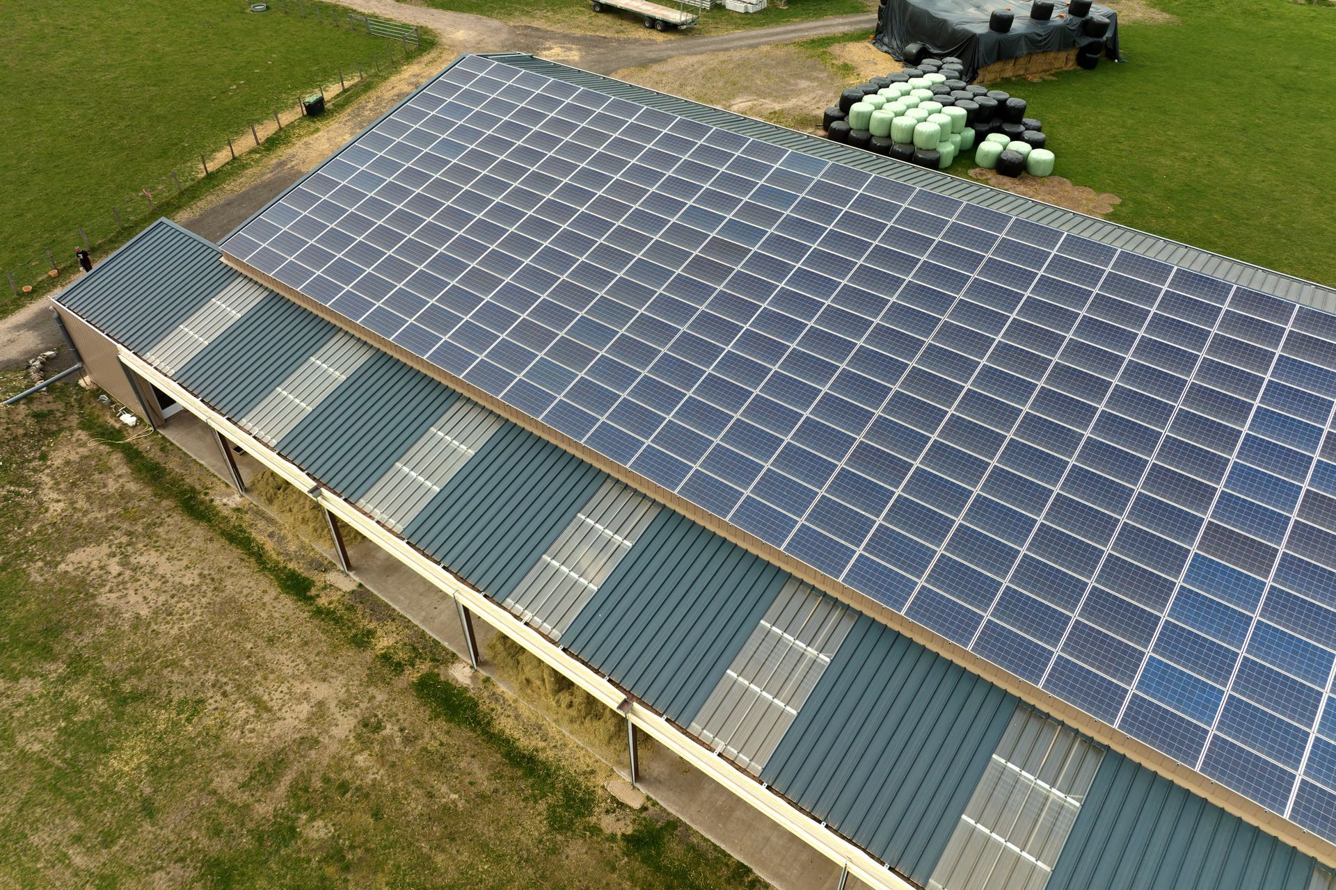 Solar panels cover the roof of a long, grey barn on a farm. Green fields surround the structure.