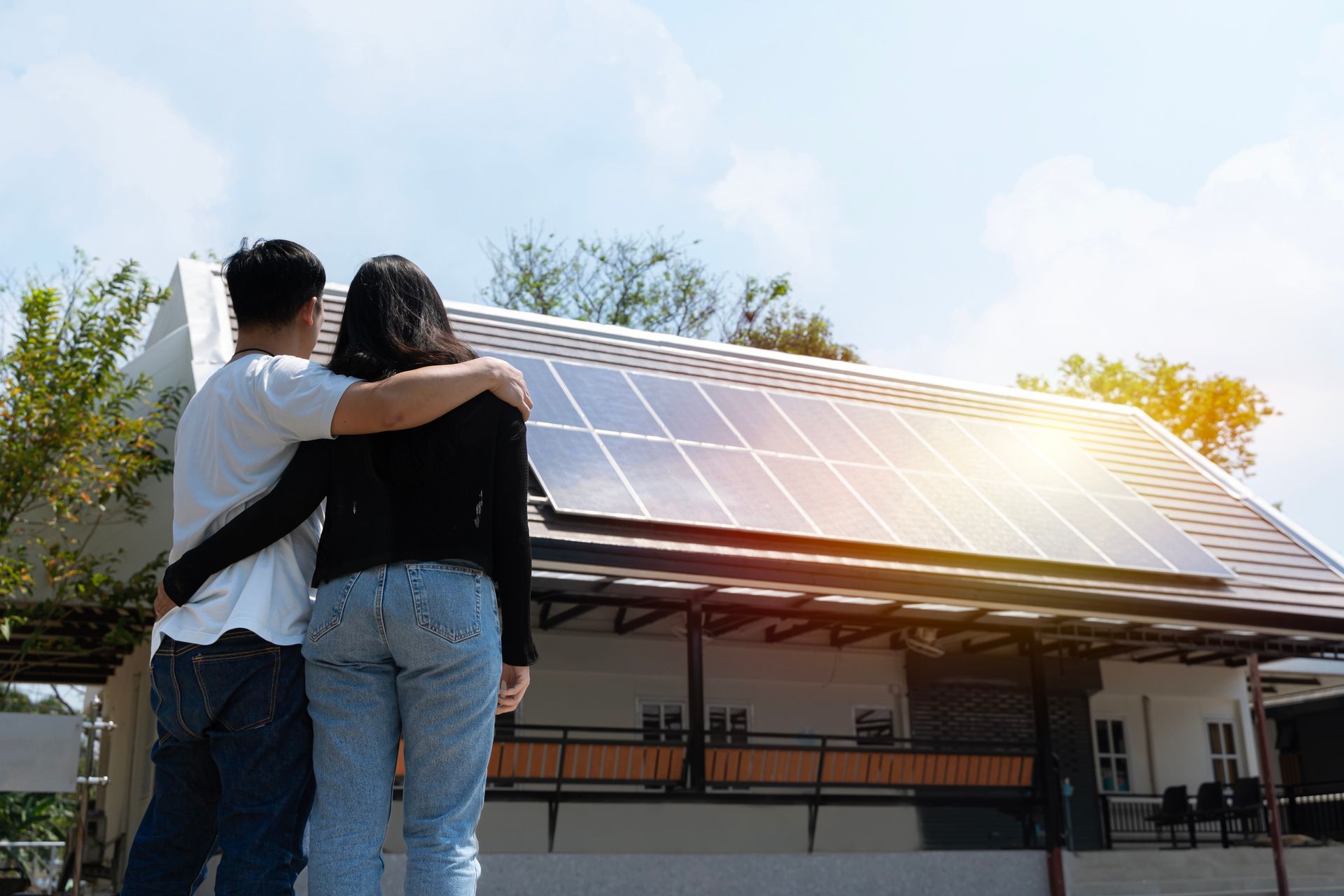 Couple with arms around each other, looking at solar panels on their house roof. Sunny day.