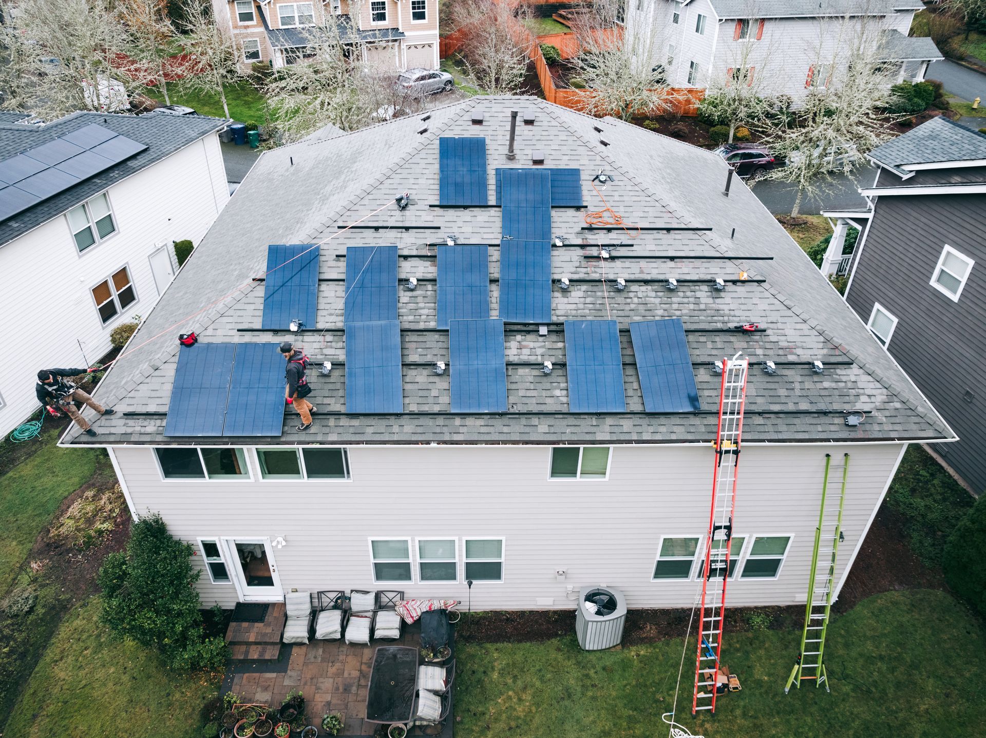 Workers installing blue solar panels on a gray roof of a light-colored house, ladders in the yard.