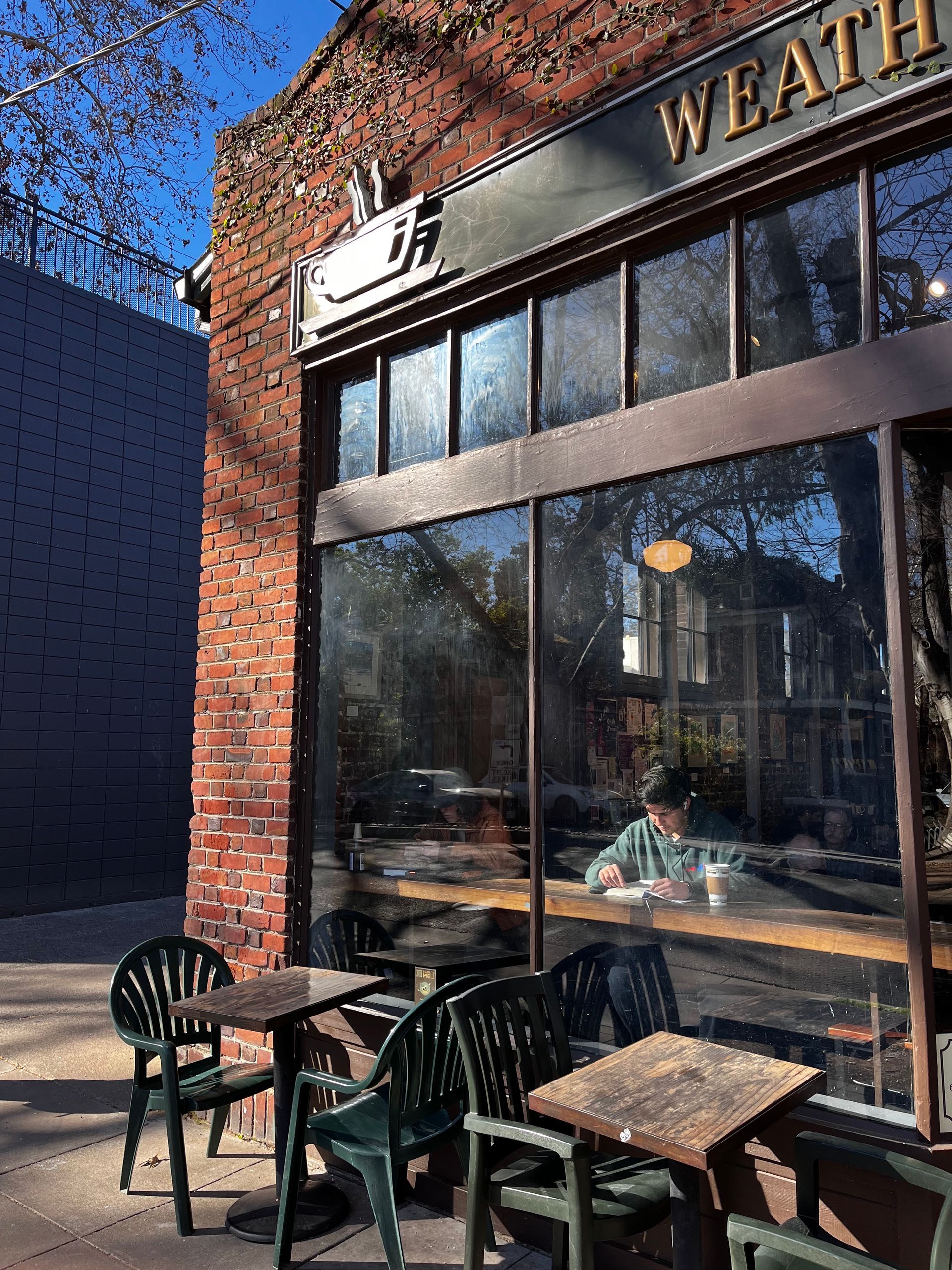 Weather coffee shop exterior with outdoor seating, brick facade, large windows, person inside, sunny day.