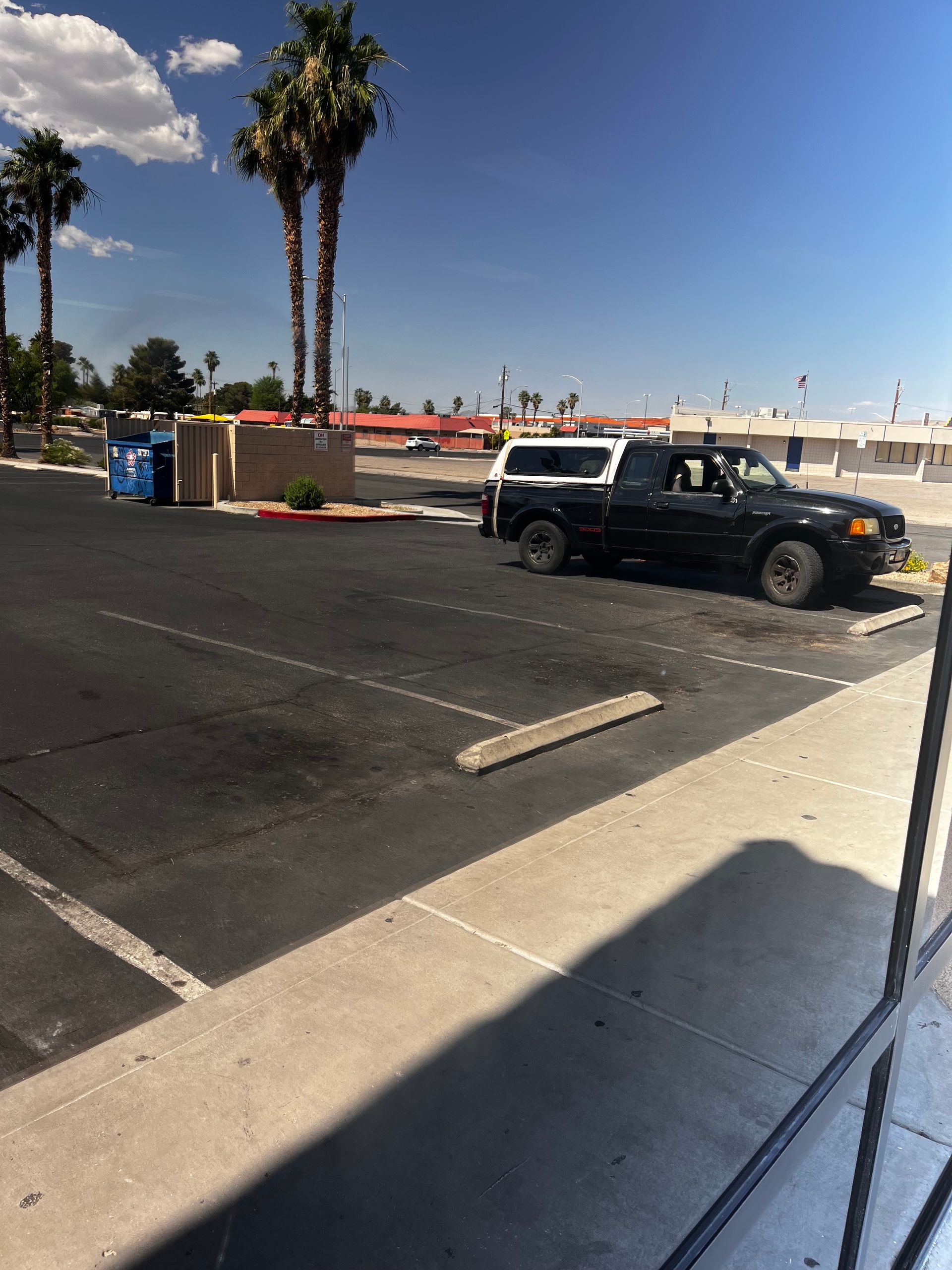 Black pickup truck parked in a mostly empty lot under a clear blue sky. Palm trees and a building are in the background.