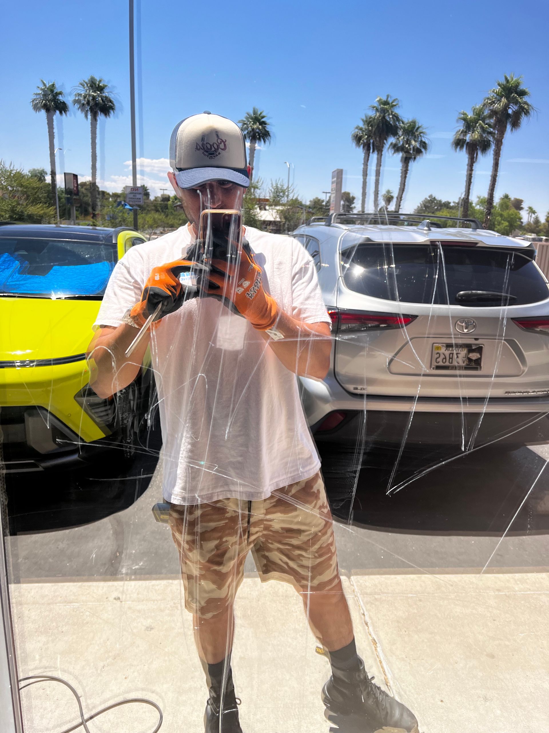 Man taking a photo reflected in glass. Wearing hat, t-shirt, camo shorts, orange gloves. Palm trees and cars in background.