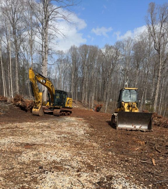 Yellow excavator and bulldozer parked on a cleared, wooded construction site under a clear blue sky.
