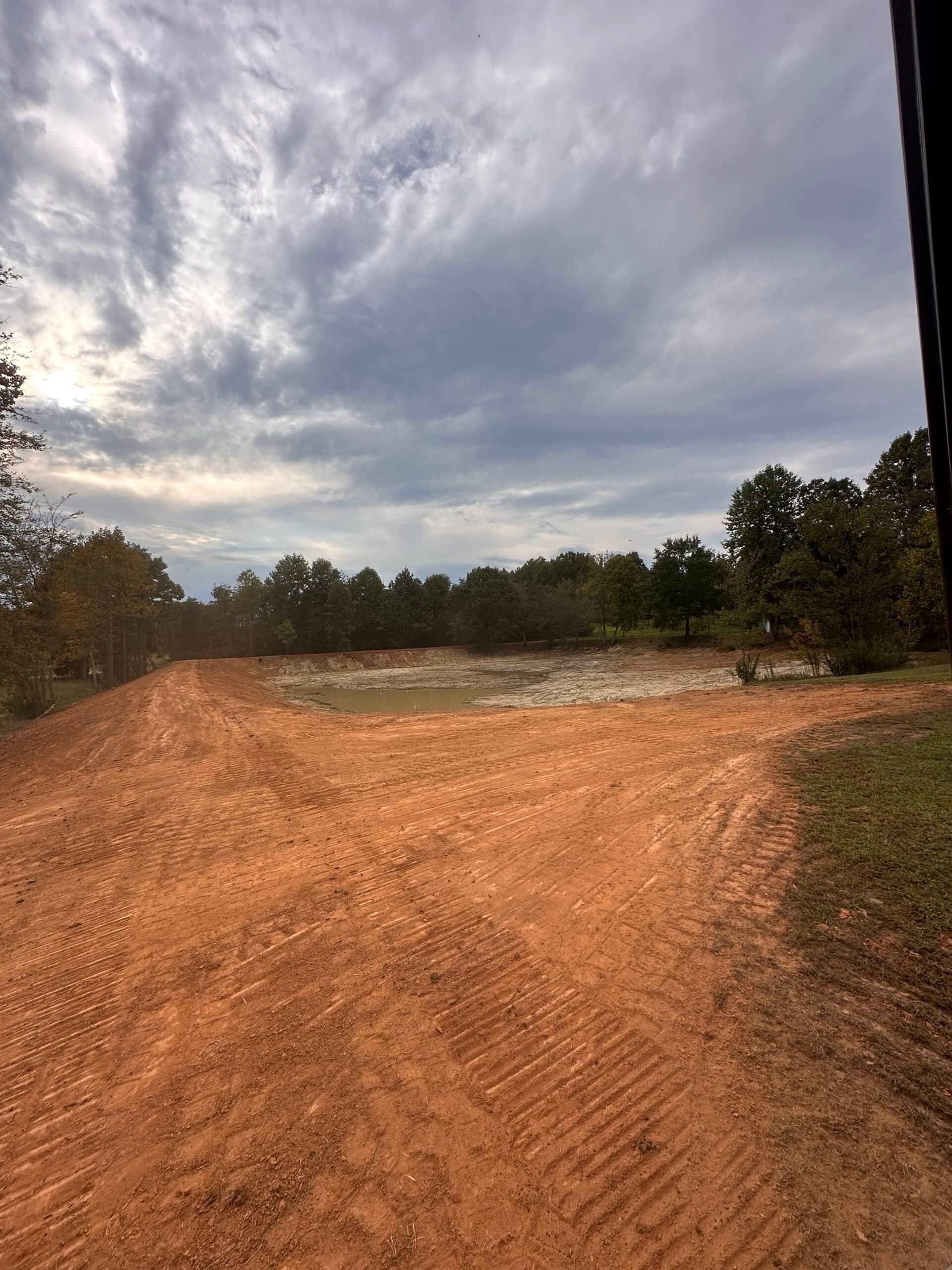 Freshly cleared red dirt slopes down toward a small pond under a cloudy sky, with trees bordering the horizon.