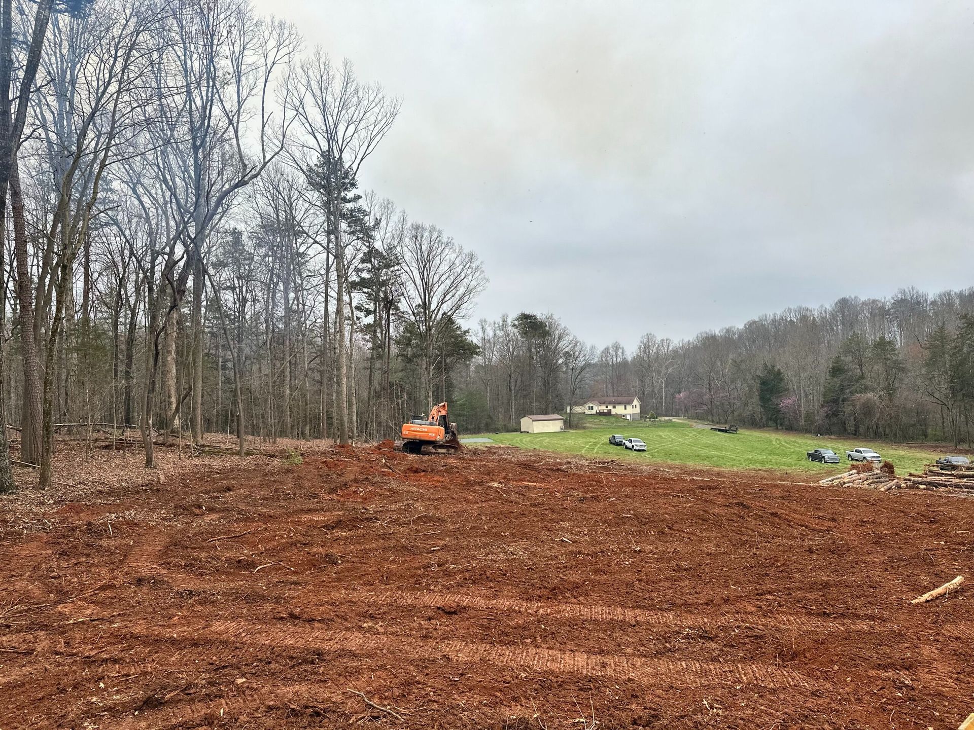 An orange construction vehicle sits on a freshly cleared patch of reddish-brown dirt, bordered by a forest and a field.