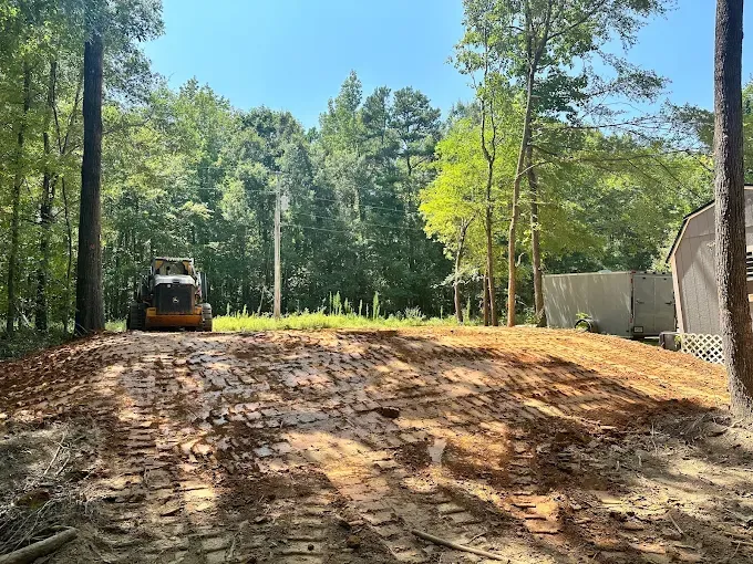 A construction vehicle sits on a freshly cleared dirt lot surrounded by trees, with a storage shed visible to the right.