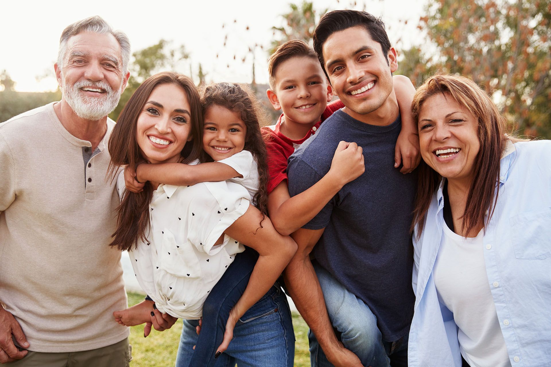 Family smiling outdoors; two adults give piggyback rides to children; grandparents smile.