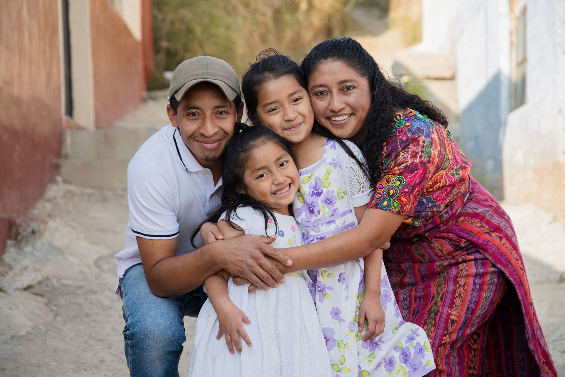 Family of four embracing outdoors; mother in colorful patterned top, children in dresses.