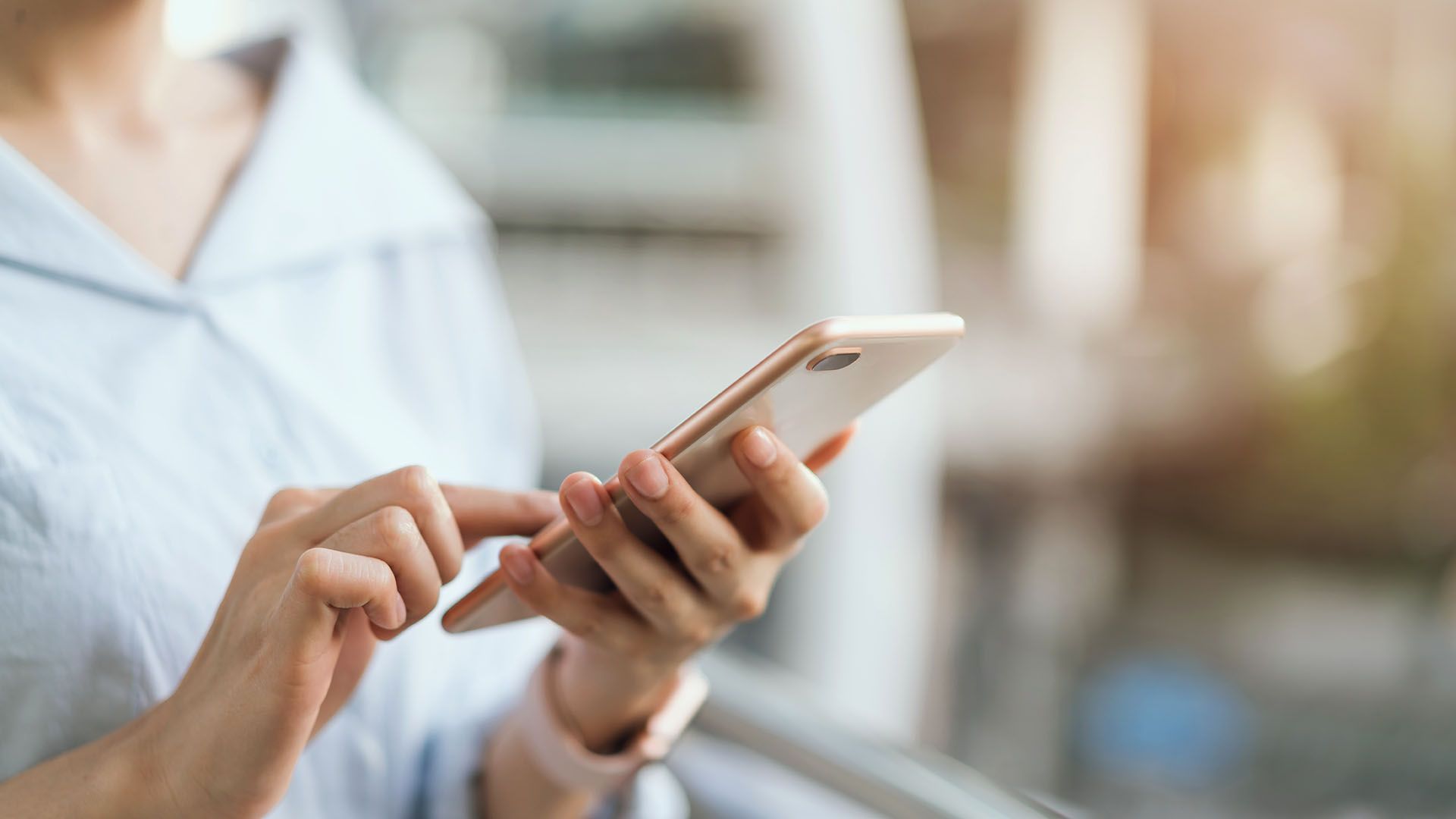 Woman in light blue shirt using a gold smartphone, outdoors, blurred background.
