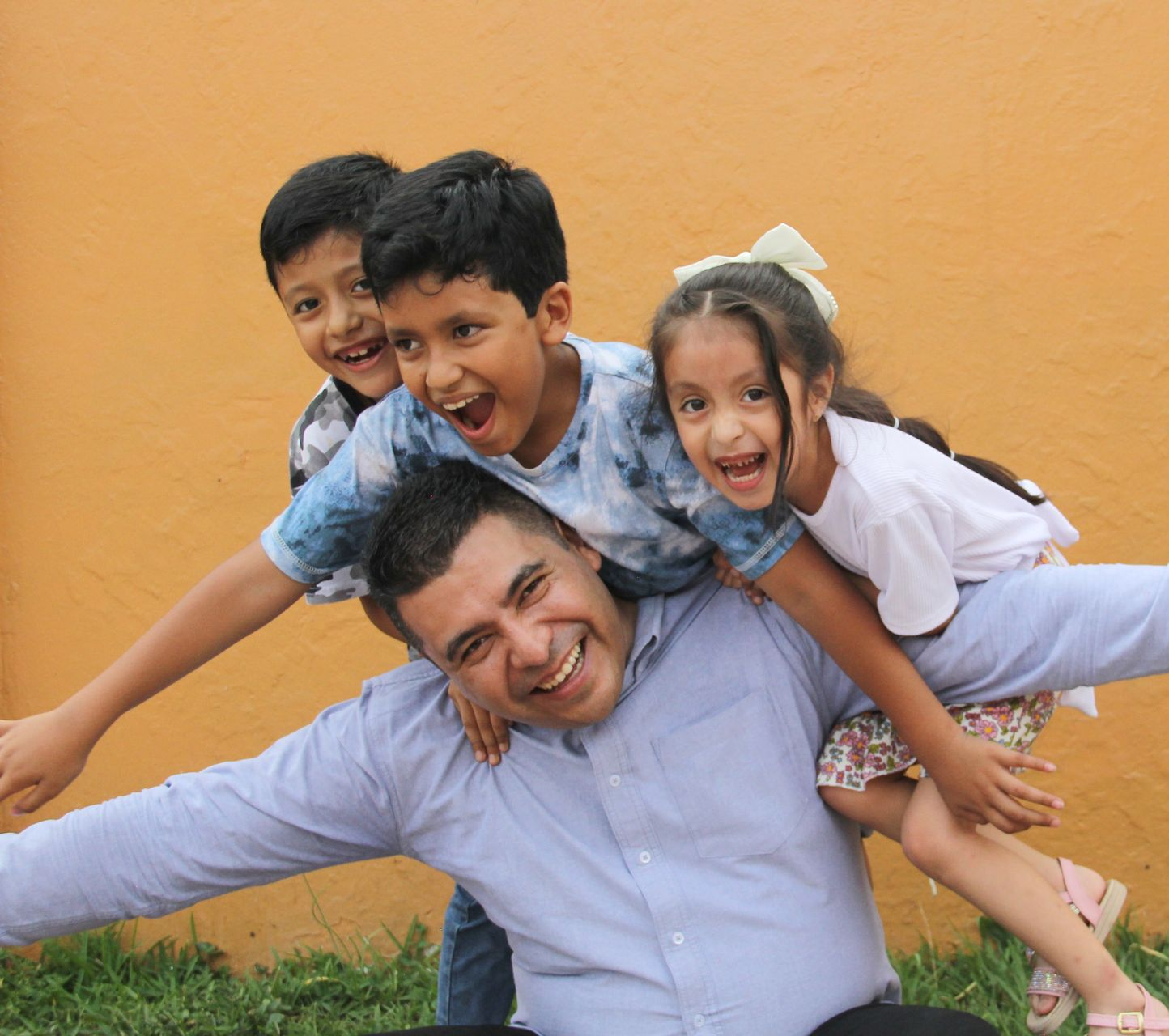 Man smiling, playing with three children, posing in front of an orange wall.
