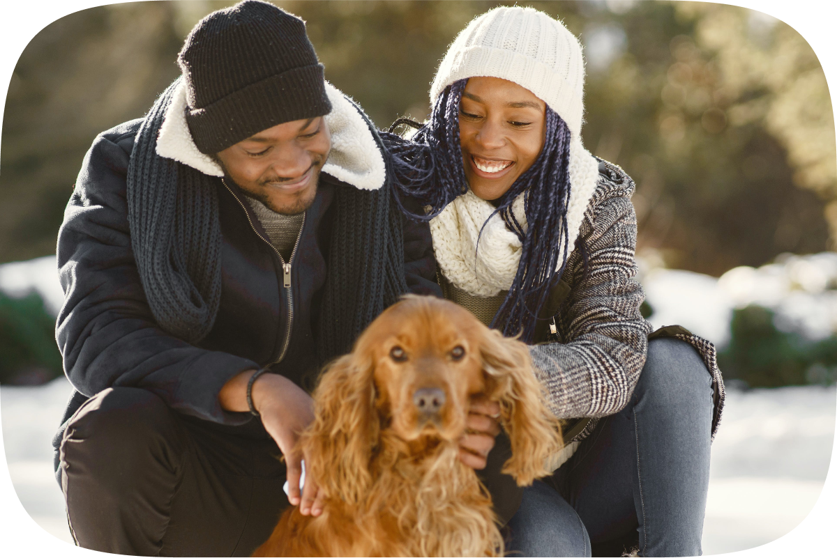 A young Black couple, dressed in winter clothes, smile and look fondly at their long haired dog as the woman pets the dog's ear