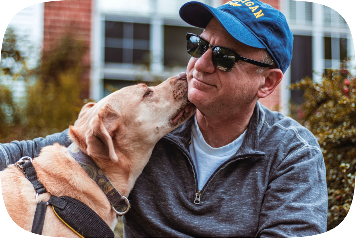 A retired white man in sunglasses and a Michigan hat lets his yellow lab give him a lick on the side of his face