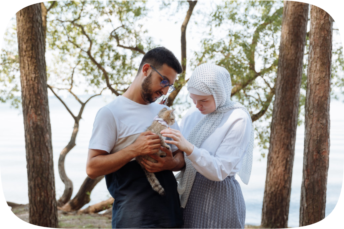 A young Muslim couple hold their kitten gently among some trees next to a lake on a sunny day