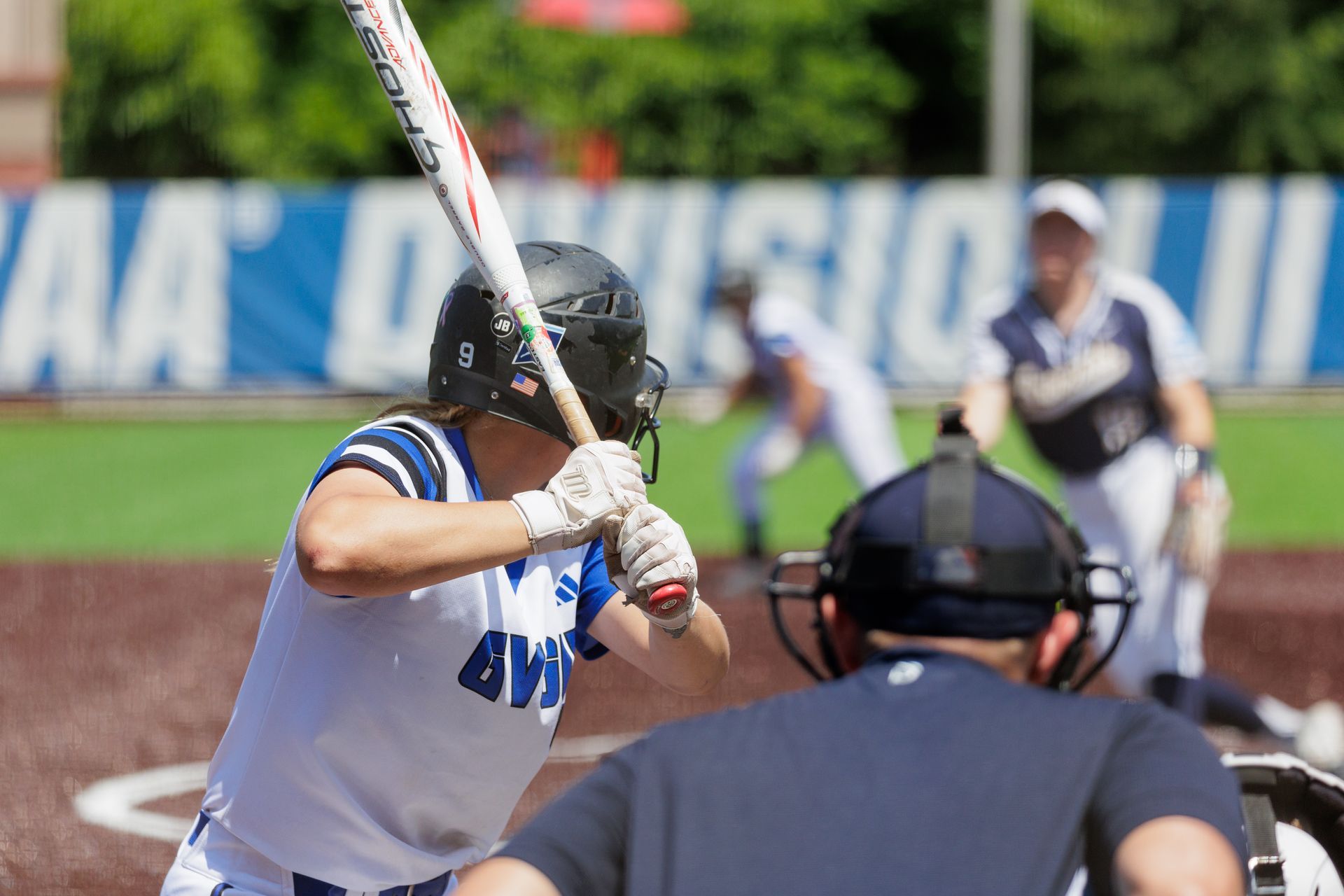 Softball batter in white and blue swings at a pitch as catcher and umpire watch behind home plate.