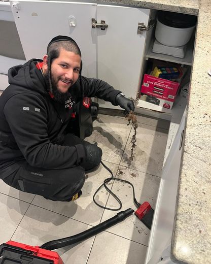 Man in black jacket and yarmulke kneels to clean under a kitchen sink with a vacuum. He smiles, looking at the camera.