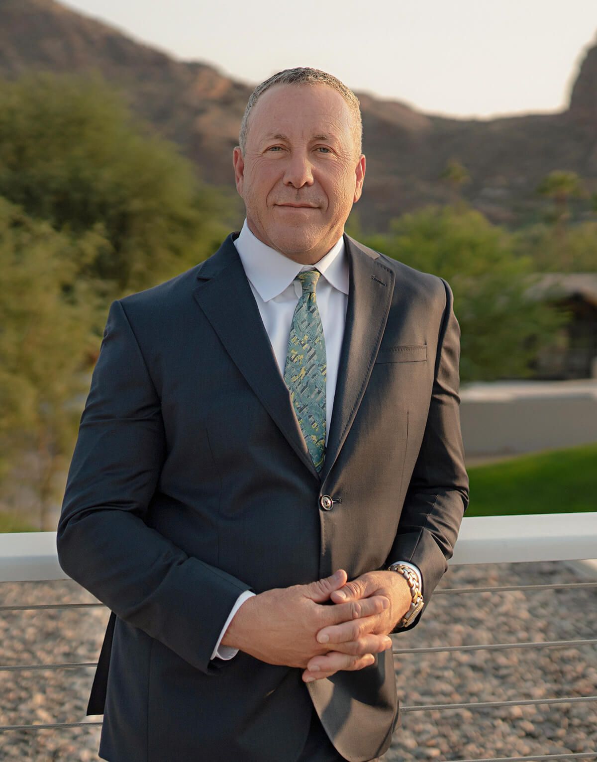 A man in a suit and tie is standing next to a railing with his hands folded.