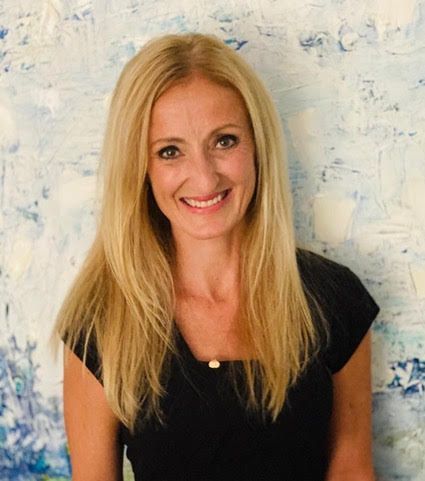 A woman with blonde hair is smiling in front of a painting.