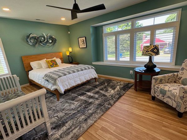 Bedroom with turquoise walls, hardwood floors, a wooden bed, a patterned rug, and a white armchair. Natural light streams in from a window.