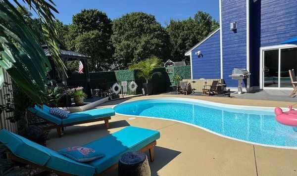 Blue pool with lounge chairs, grill, and blue house in a sunny backyard.