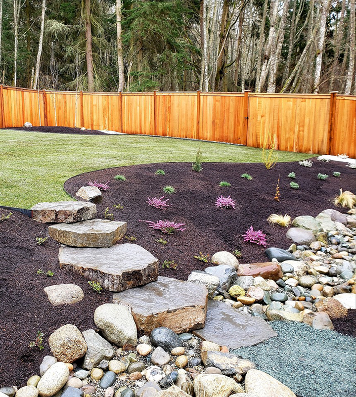 a wooden fence surrounds a lush green yard with rocks and flowers .