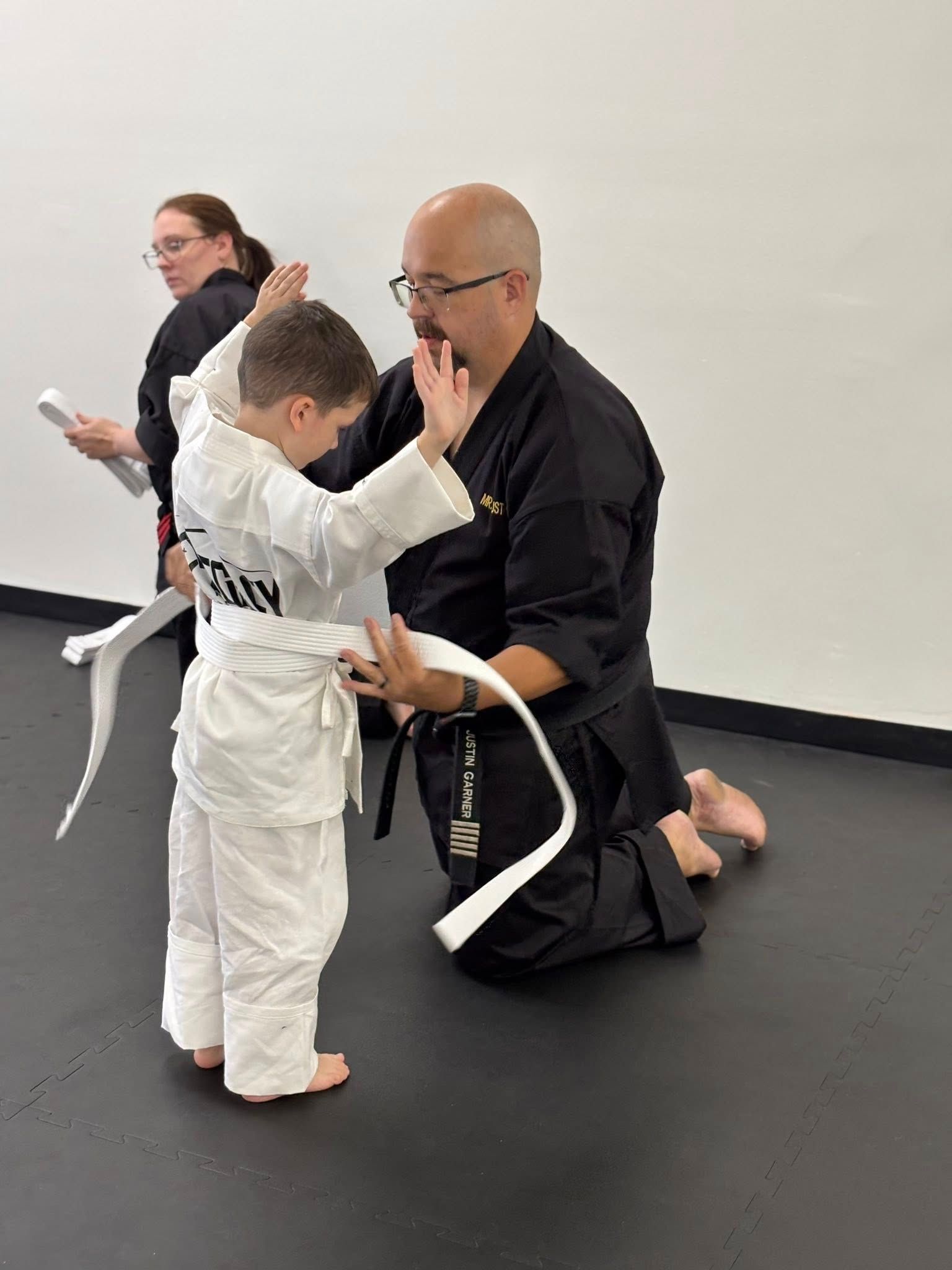 Man ties white belt on a young child in a dojo; another person watches.