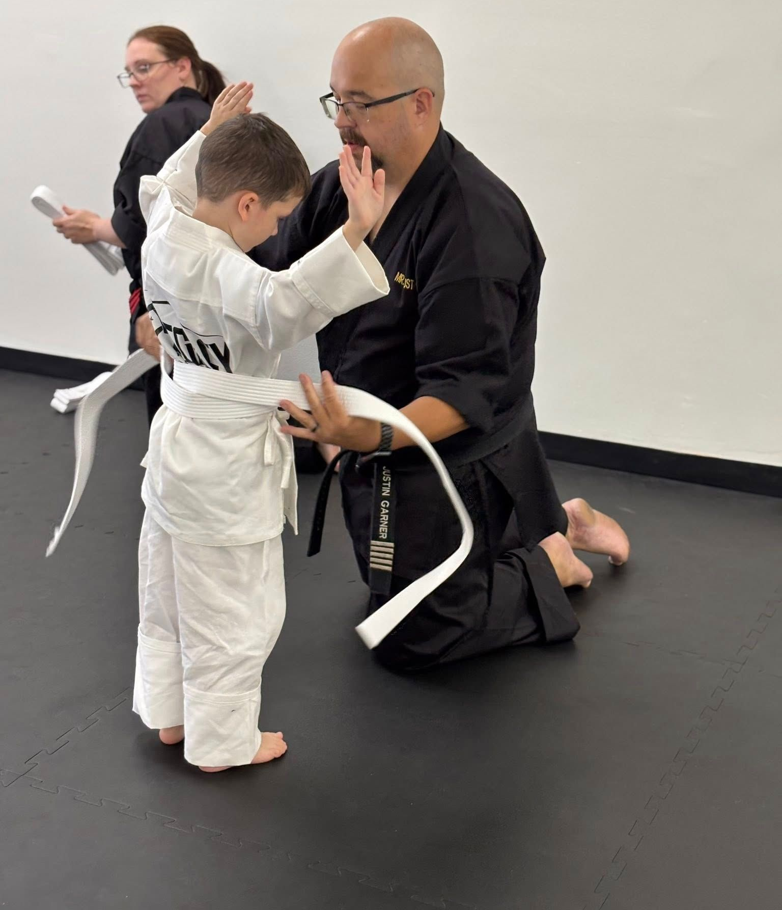 Instructor helping a child tie a white karate belt in a dojo.