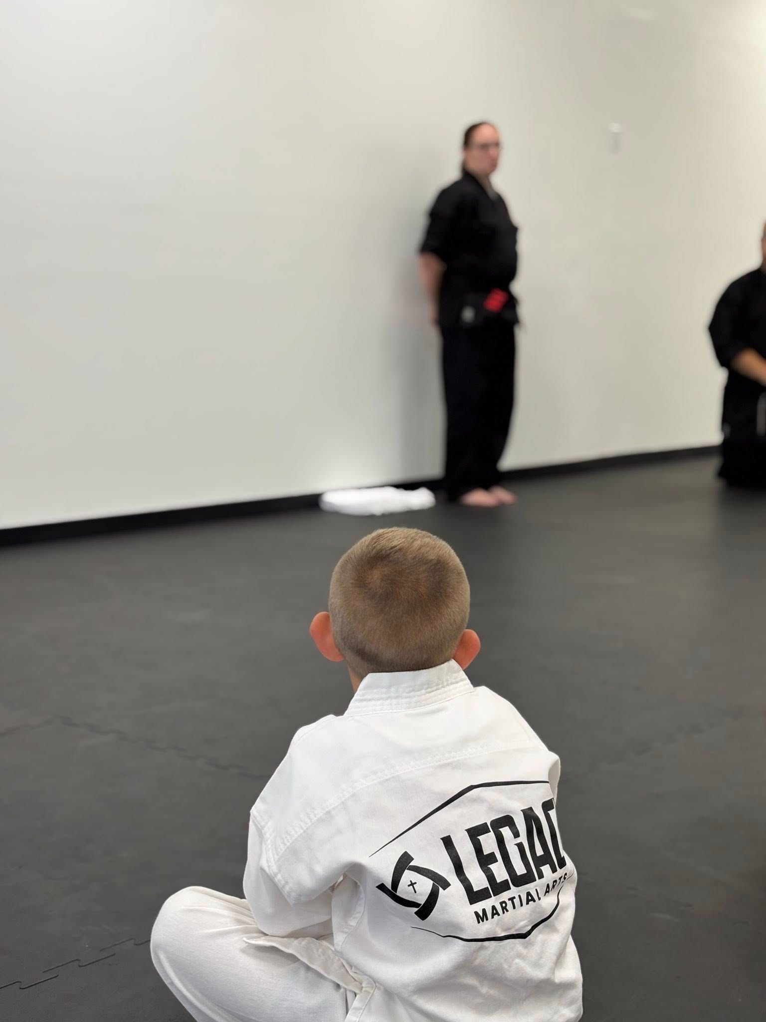Child in white gi sits on the floor, facing instructor in black uniform in a dojo.