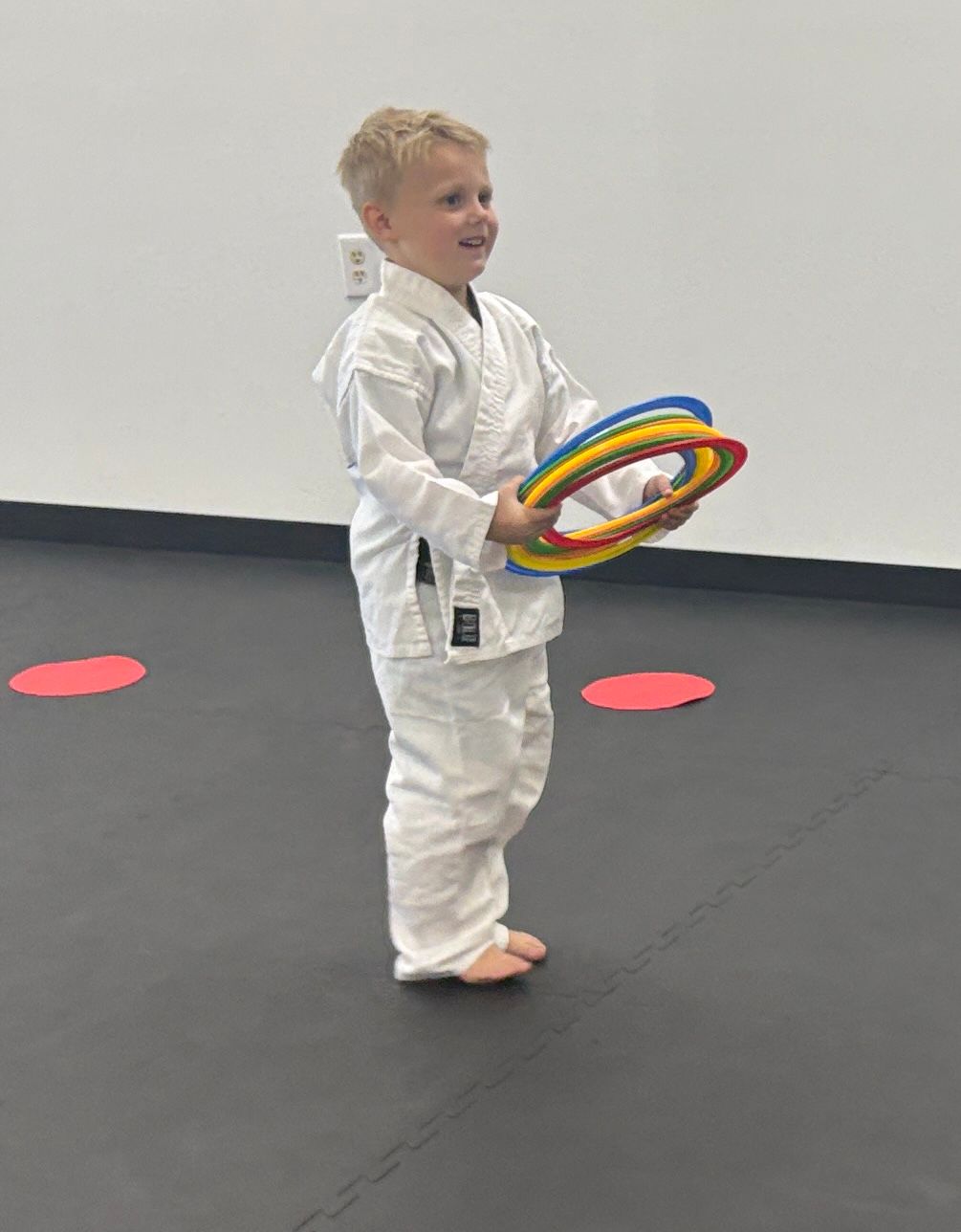 Young child in a white martial arts uniform holding colorful rings, smiling, on a black floor with red dots.