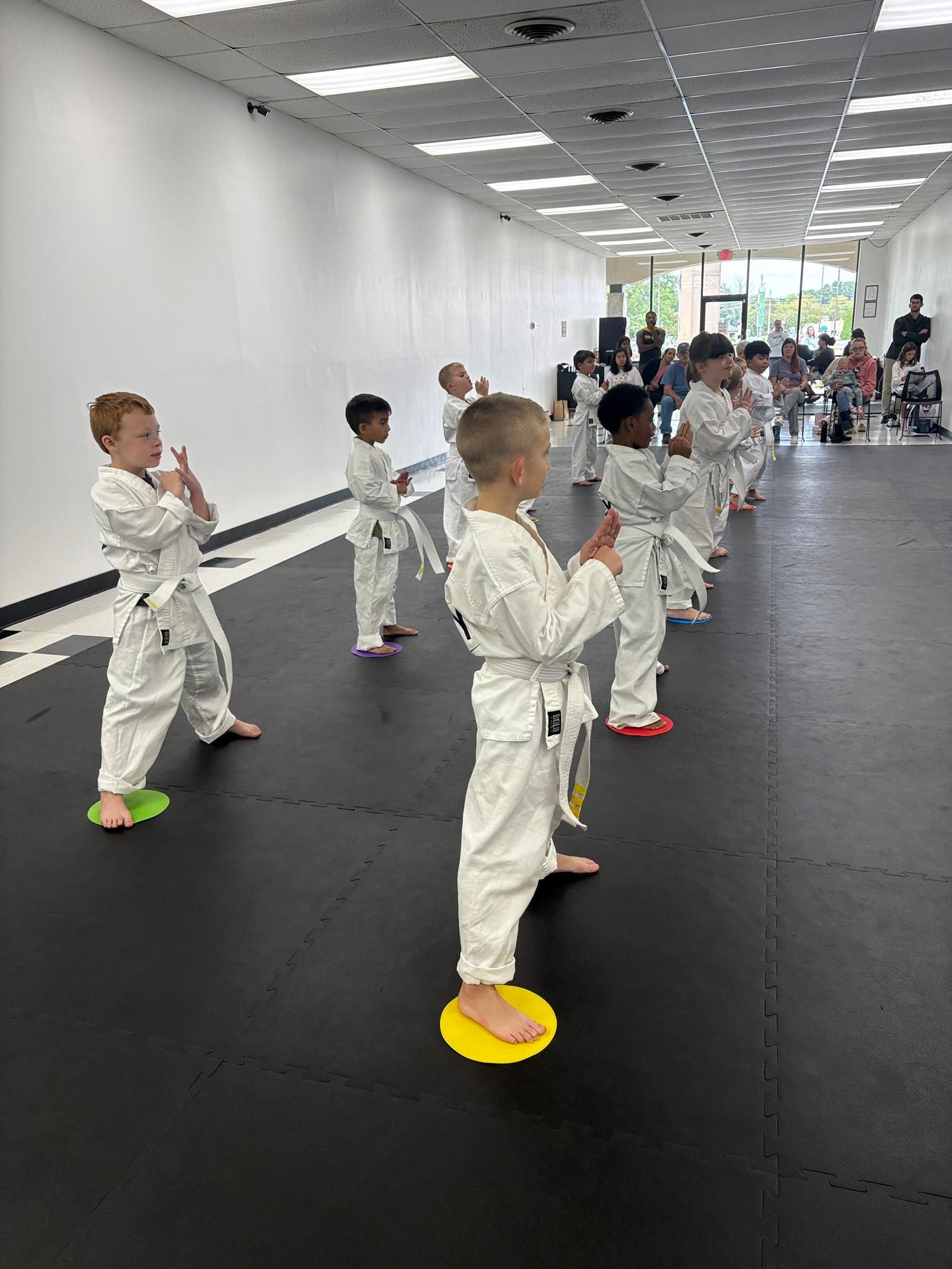 Children in karate uniforms practice in a dojo, standing on colored markers.