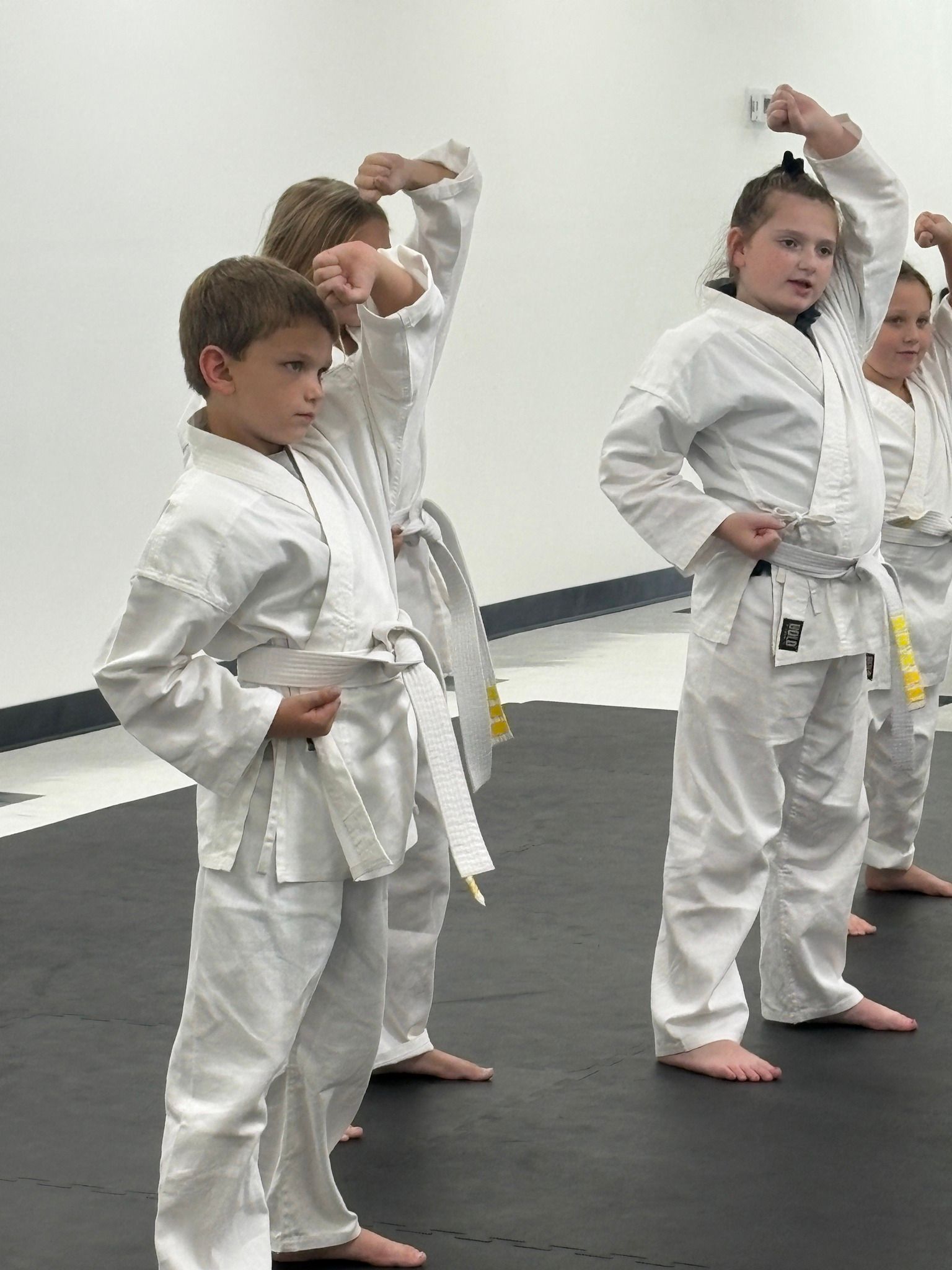 Children in white karate uniforms practicing a move, standing on a dark mat.